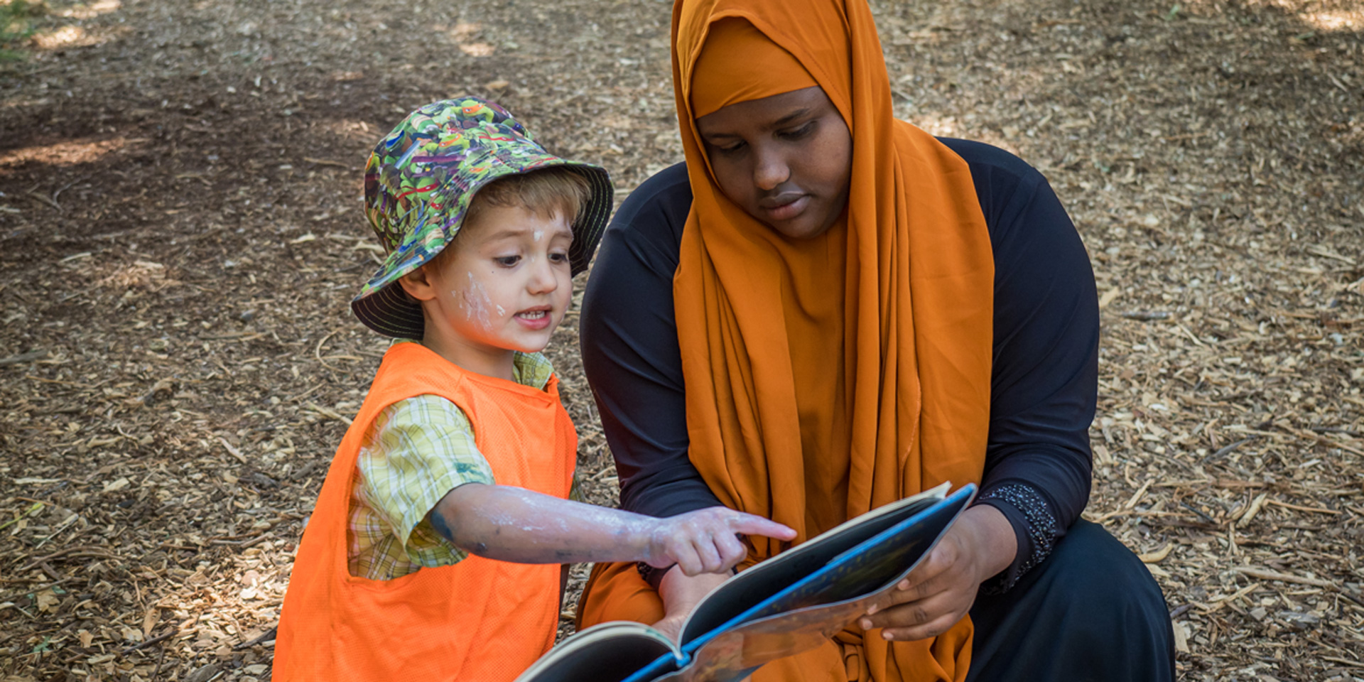 A woman reads a book outdoors with a small boy who has paint and dirt on his face.