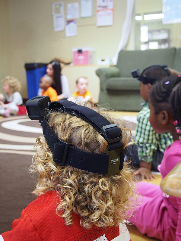 A child wearing a GoPro on their head sits in a preschool circle with other children.