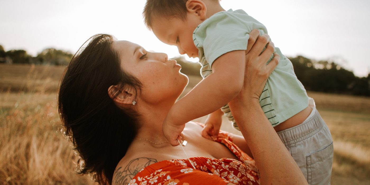 A woman making a kissing face at a baby she's holding up, with a sun setting behind them