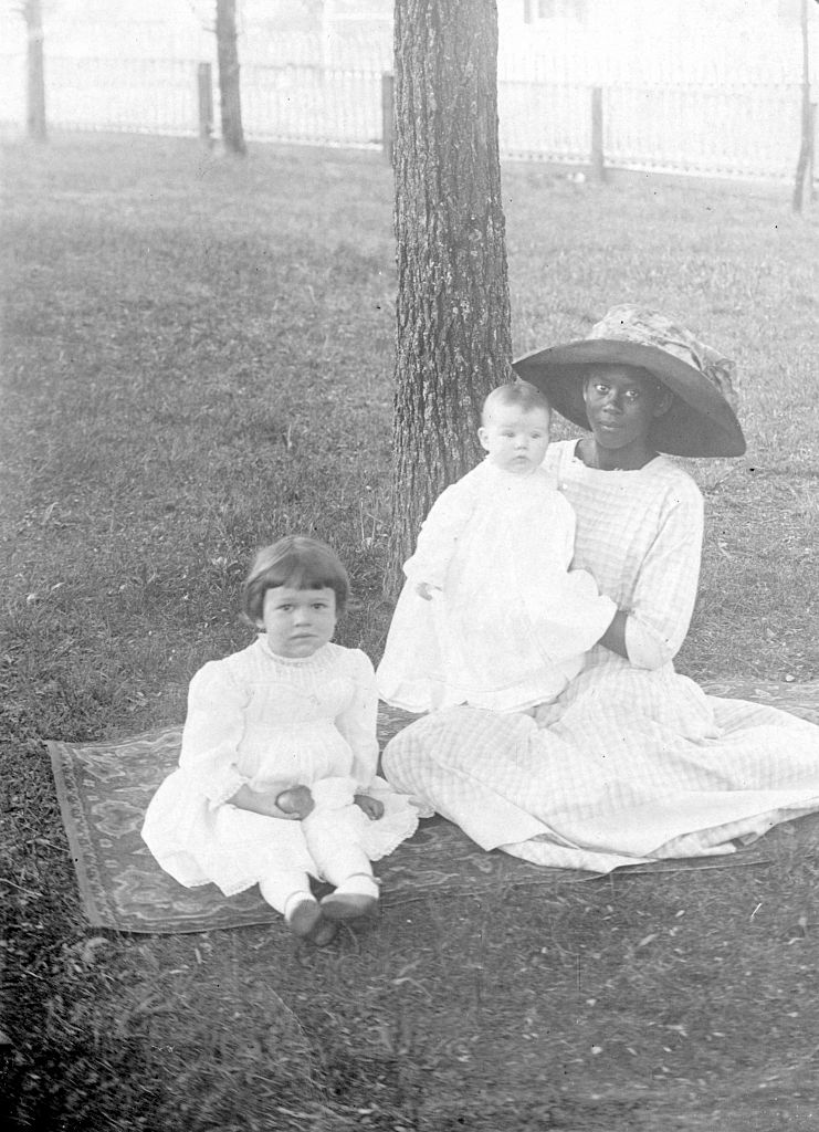 An African American nanny is wearing a light-colored dress and a large hat, she is sitting on a rug on the grass holding a Caucasian baby and sitting next to a Caucasian young girl, they are all sitting in front of a tree, 1920. (Photo by JHU Sheridan Libraries/Gado/Getty Images).