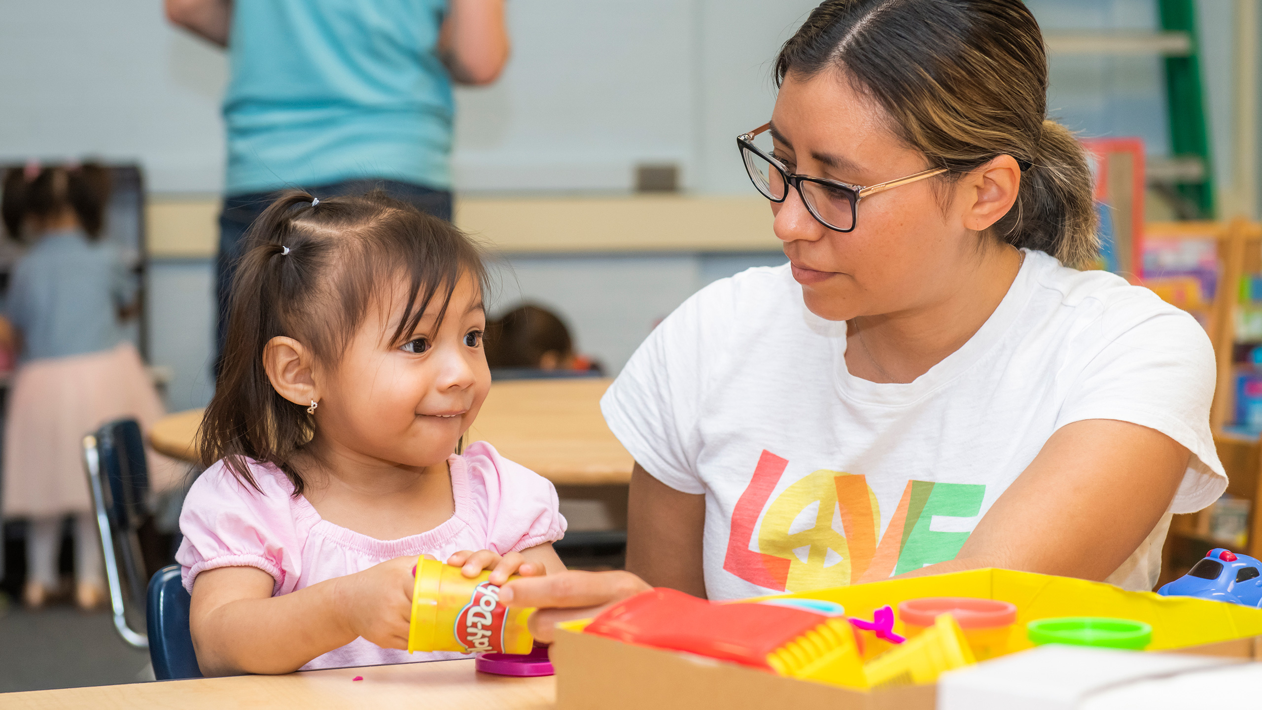 An image of a woman with glasses sitting at a child's table with a smiling kid playing with Play-Dough